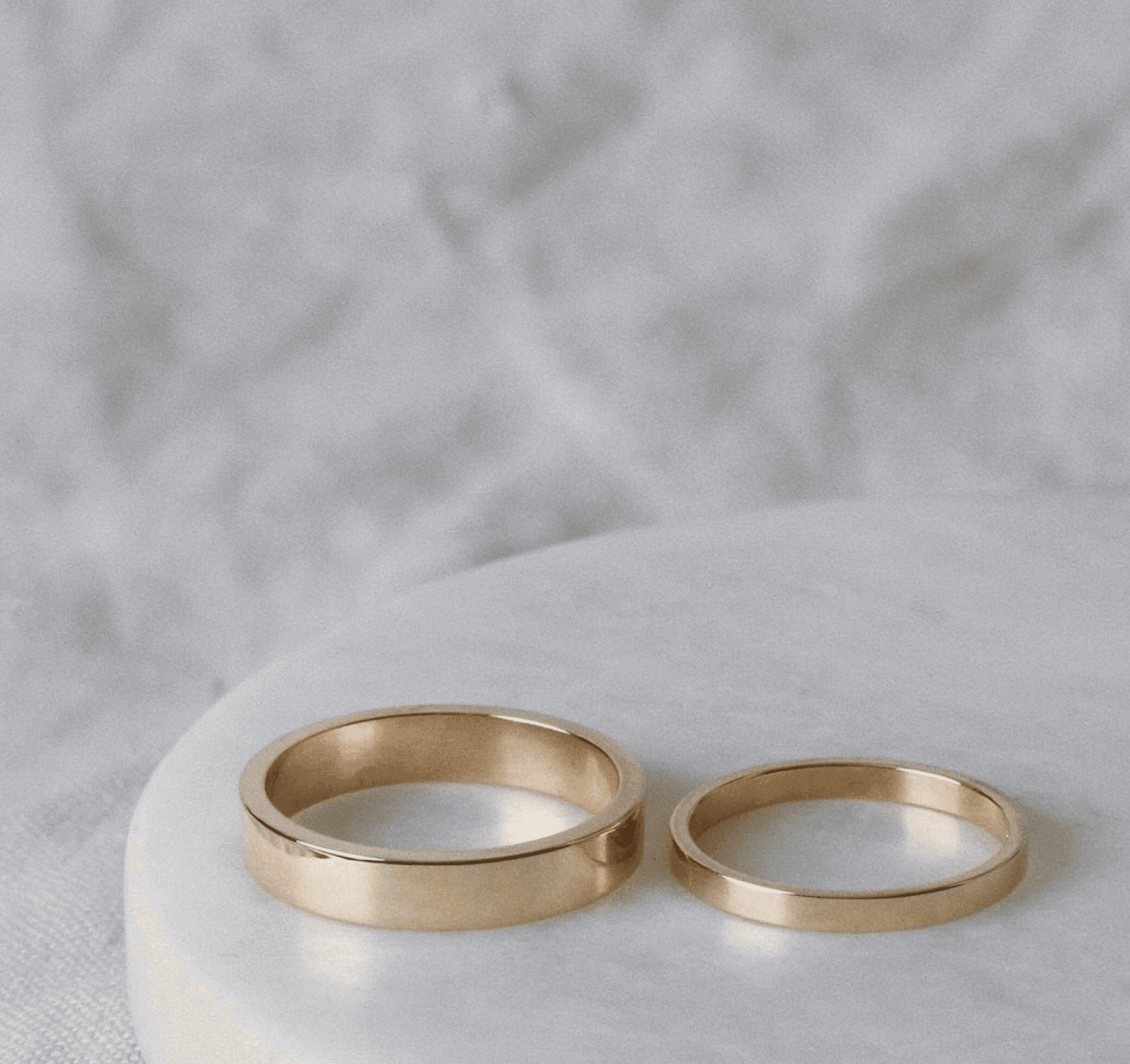 Pair of plain polished 18ct yellow gold wedding bands in two widths, resting side by side on a white marble disc against draped white linen