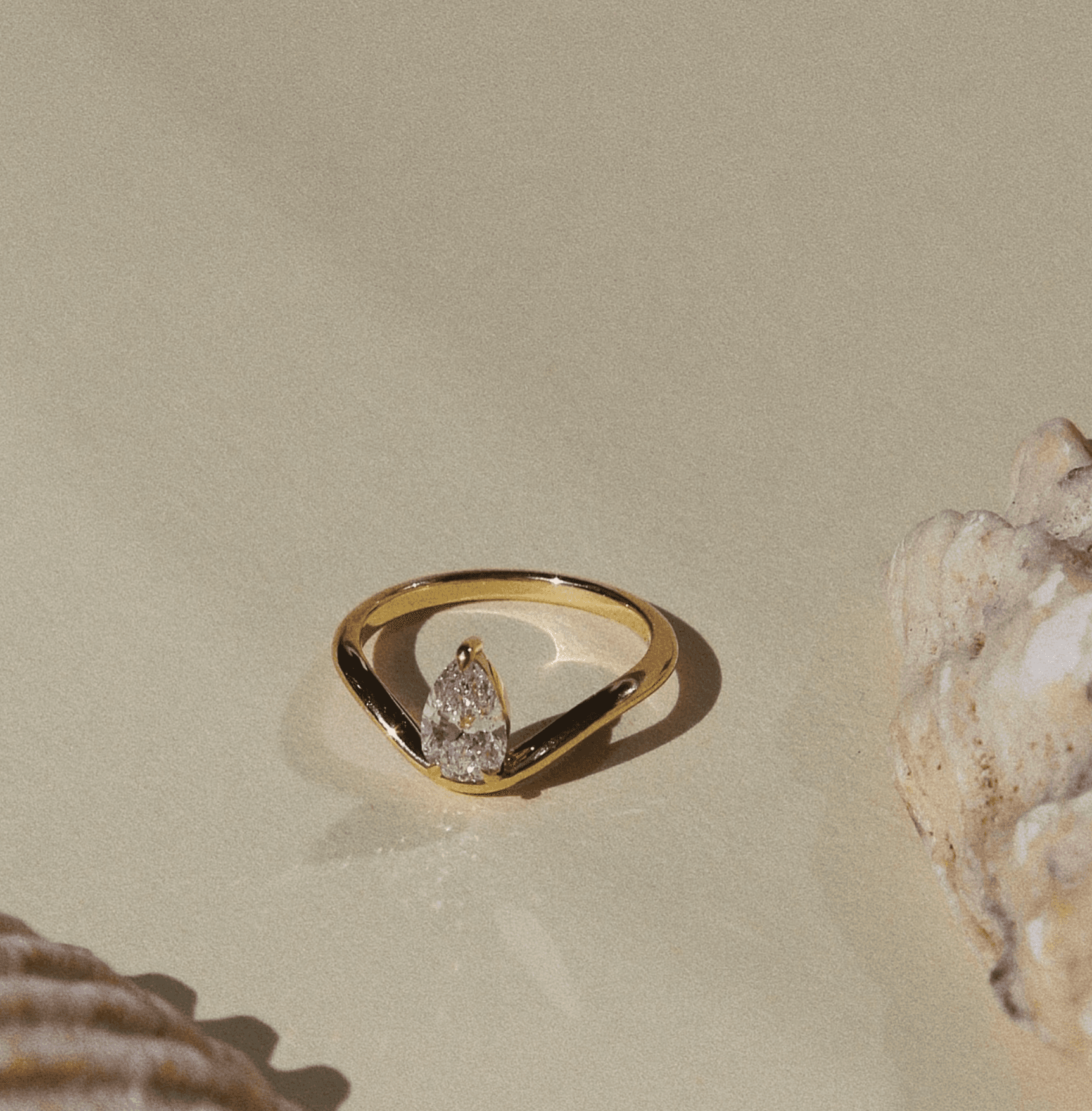 Pear-cut diamond engagement ring with sculptural chevron band, cast in 18ct yellow gold, photographed on warm sand with sea shells